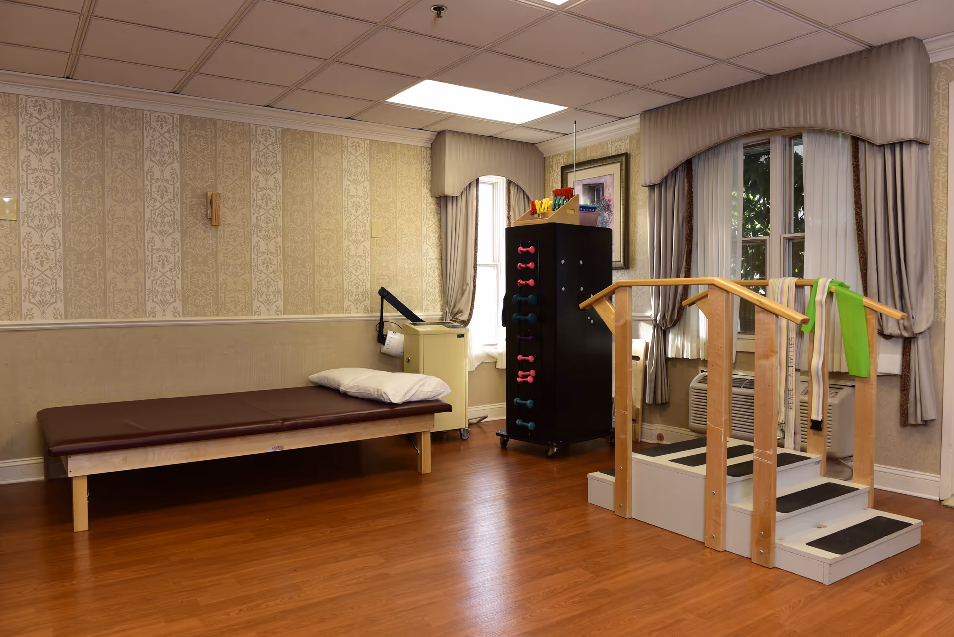 A therapy room with a padded examination table, a set of stairs with handrails for physical therapy, resistance bands hanging on the rails, a rack with colorful dumbbells, and windows with curtains letting in natural light.