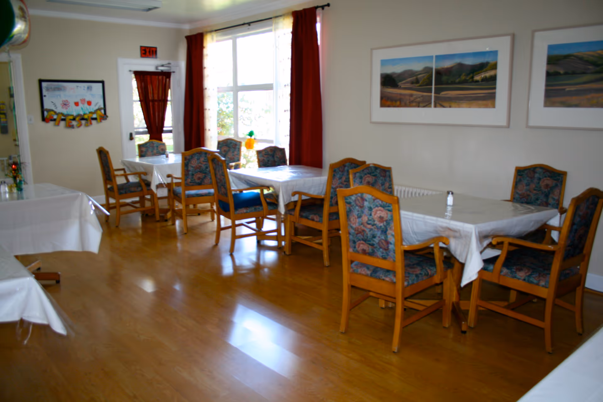Dining area with wooden tables and patterned chairs in a senior living facility.