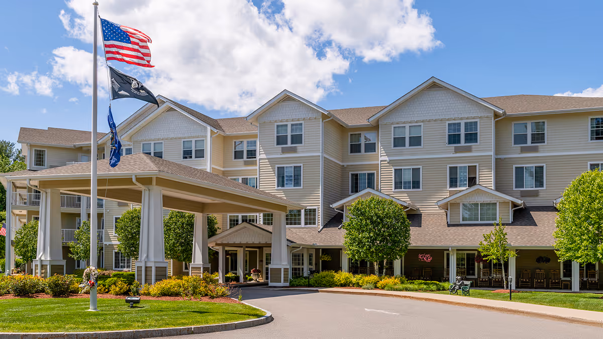 Exterior view of a multi-story senior living facility named Holiday Birch Heights with a covered entrance, well-maintained landscaping, and three flags flying on a flagpole under a partly cloudy sky.