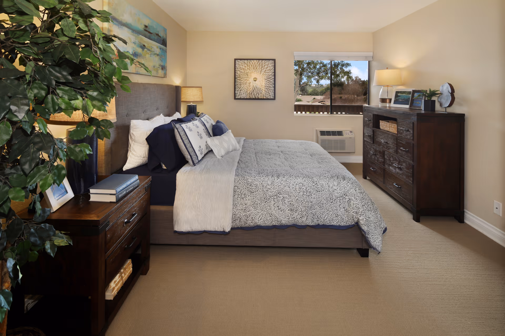 A neatly arranged bedroom with a large bed featuring patterned bedding and multiple pillows. On the left side, there is a wooden nightstand with books and a lamp, next to a large leafy plant. On the right side, there is a dark wooden dresser with framed photos, a lamp, and decorative items. A window shows a view of trees and rooftops outside. The walls are painted beige and the carpet is light-colored.