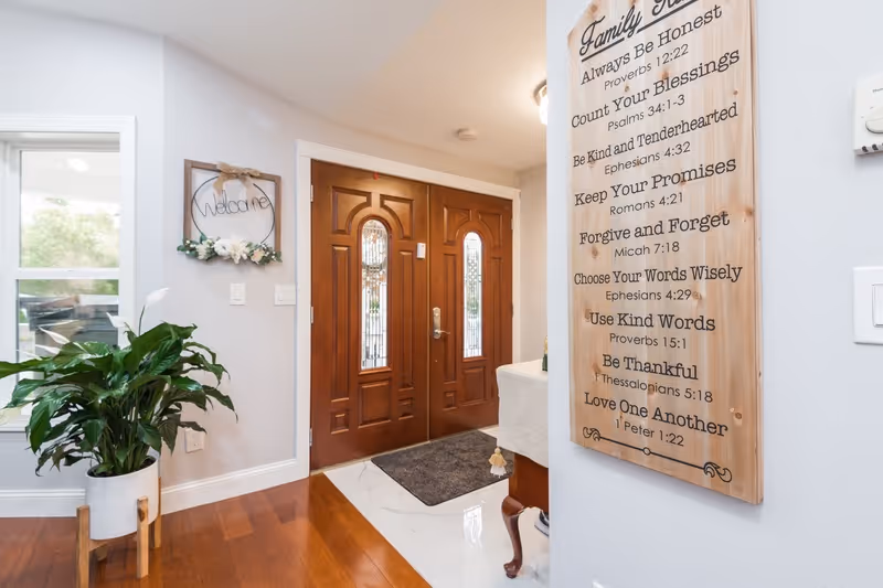 Bright entryway with double wooden front doors, a potted plant, a welcome sign, and a wooden wall plaque listing family rules.