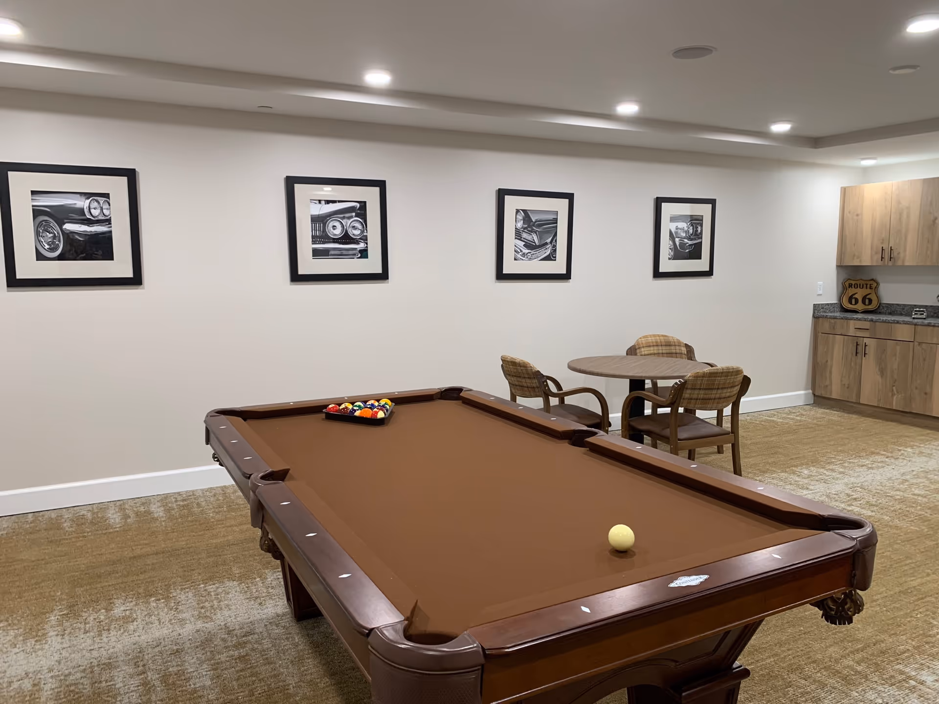 A recreational room with a brown pool table in the foreground, a round table with three plaid upholstered chairs, and a kitchenette area with wooden cabinets and a Route 66 sign. Four black and white framed photographs of classic car details hang on the white wall.
