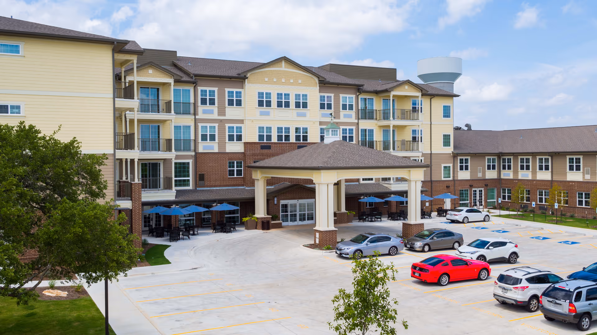 Exterior view of a senior living facility with a large covered entrance, multiple floors with balconies, outdoor seating areas with blue umbrellas, a parking lot with several cars, and a water tower in the background under a partly cloudy sky.