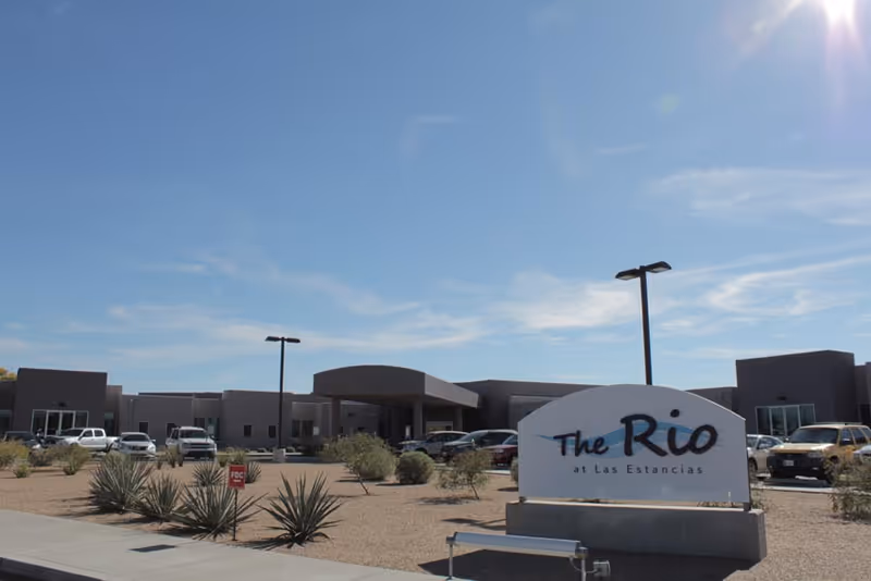 Exterior view of a single-story building with a parking lot in front under a clear blue sky. A sign in the foreground reads 'The Rio at Las Estancias'. Desert landscaping with small bushes and agave plants surrounds the area.
