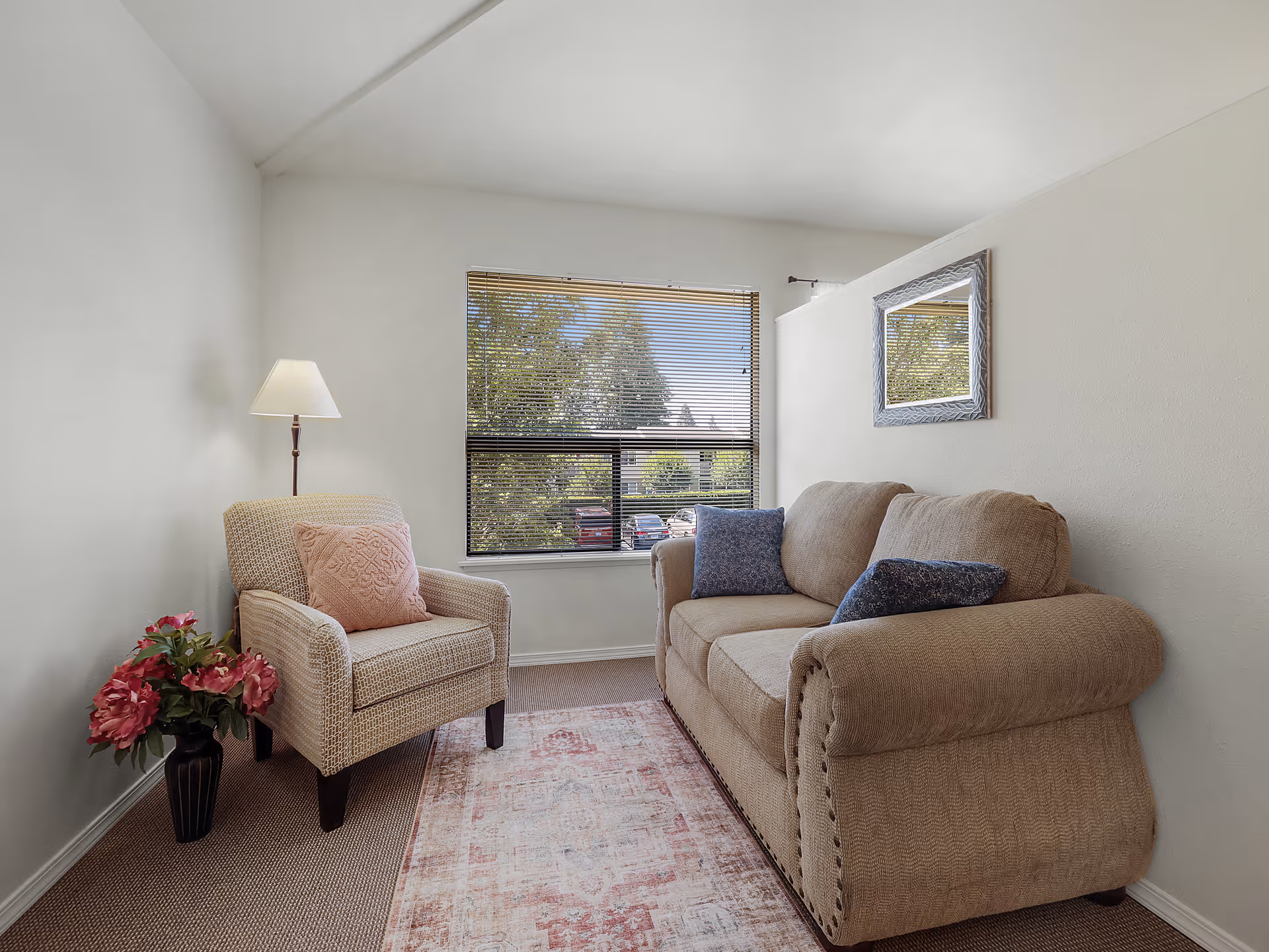 A cozy living room with a beige sofa adorned with two blue pillows, a beige armchair with a pink cushion, a floor lamp, a vase with pink flowers, a window with blinds showing trees and parked cars outside, and a decorative mirror on the wall.