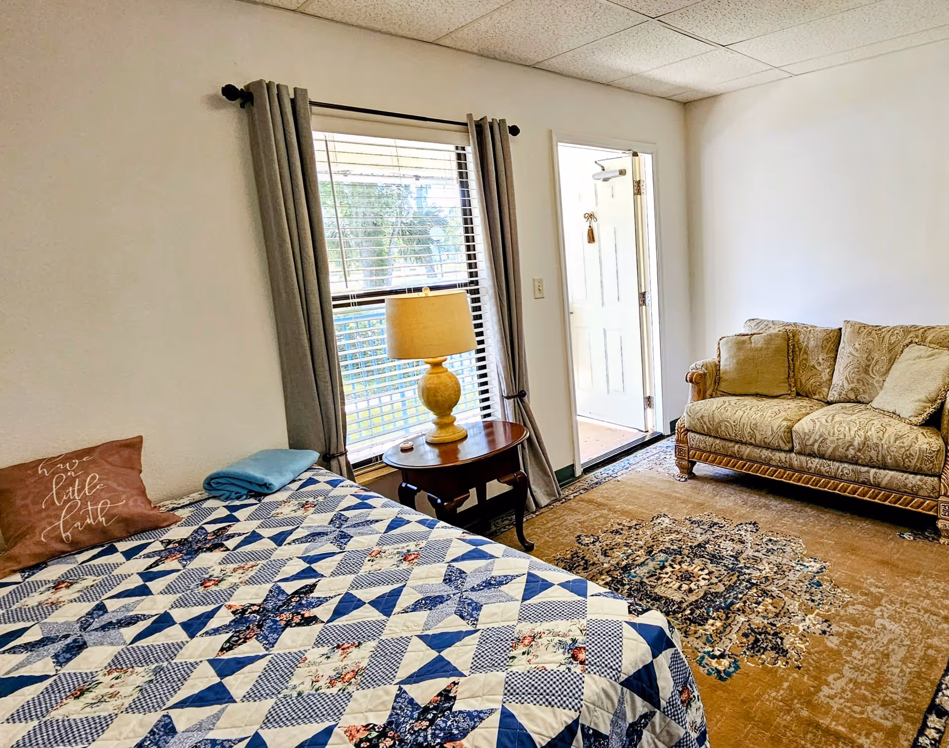 A cozy room with a bed covered in a blue and white patterned quilt, a brown pillow with the words 'have a little faith', and a folded blue blanket. Next to the bed is a wooden side table with a yellow lamp. There is a window with gray curtains behind the table. Across from the bed is a beige patterned sofa on a large decorative rug. An open door leads outside, letting in natural light.