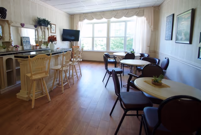 A bright dining area with wooden flooring featuring round tables with chairs and a counter with high stools. Large windows with curtains allow natural light to fill the room. The walls are decorated with framed pictures and a small TV is mounted near the window.