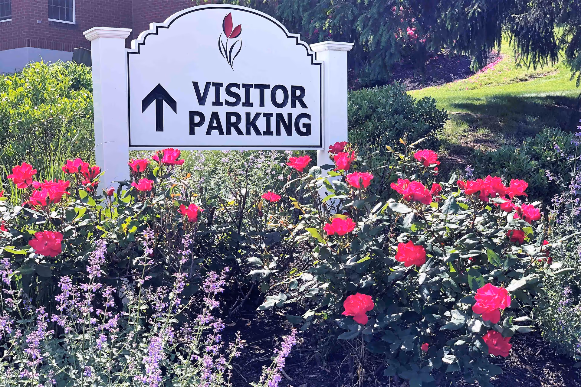 A white sign with a red tulip logo and black text reading 'VISITOR PARKING' with an upward arrow, surrounded by blooming pink and purple flowers and green shrubbery in a garden area.