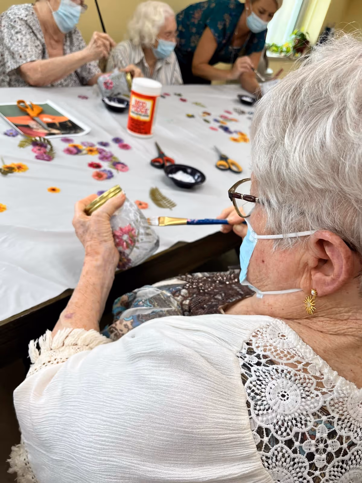 A group of elderly women wearing face masks are seated around a table engaged in a craft activity involving painting and decorating glass containers with floral designs. Various craft supplies like scissors, glue, and decorative pieces are spread out on the table.