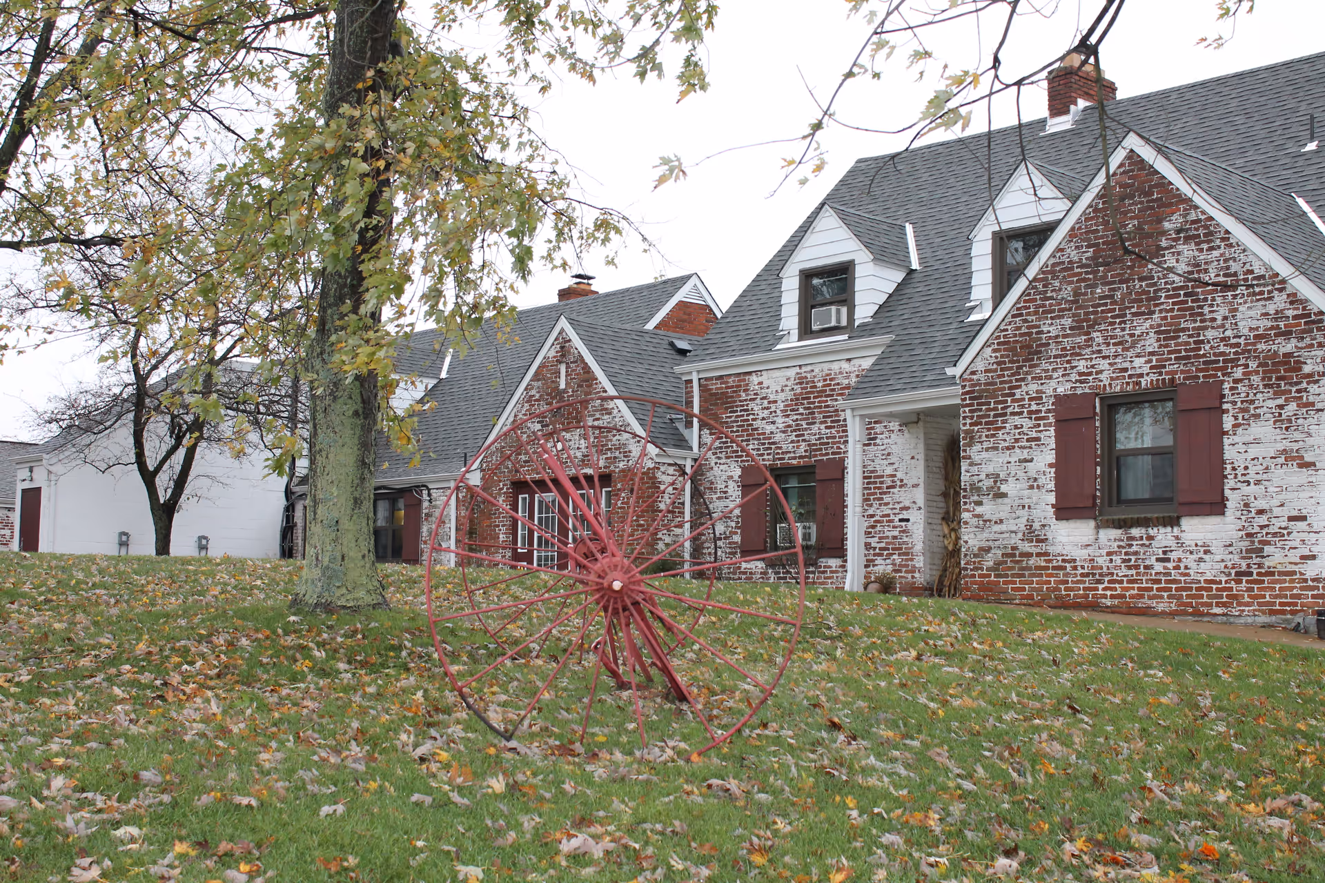 Whitewashed red brick, residential-style building with dormer windows seen from a grassy front yard featuring a decorative red metal wheel and trees.
