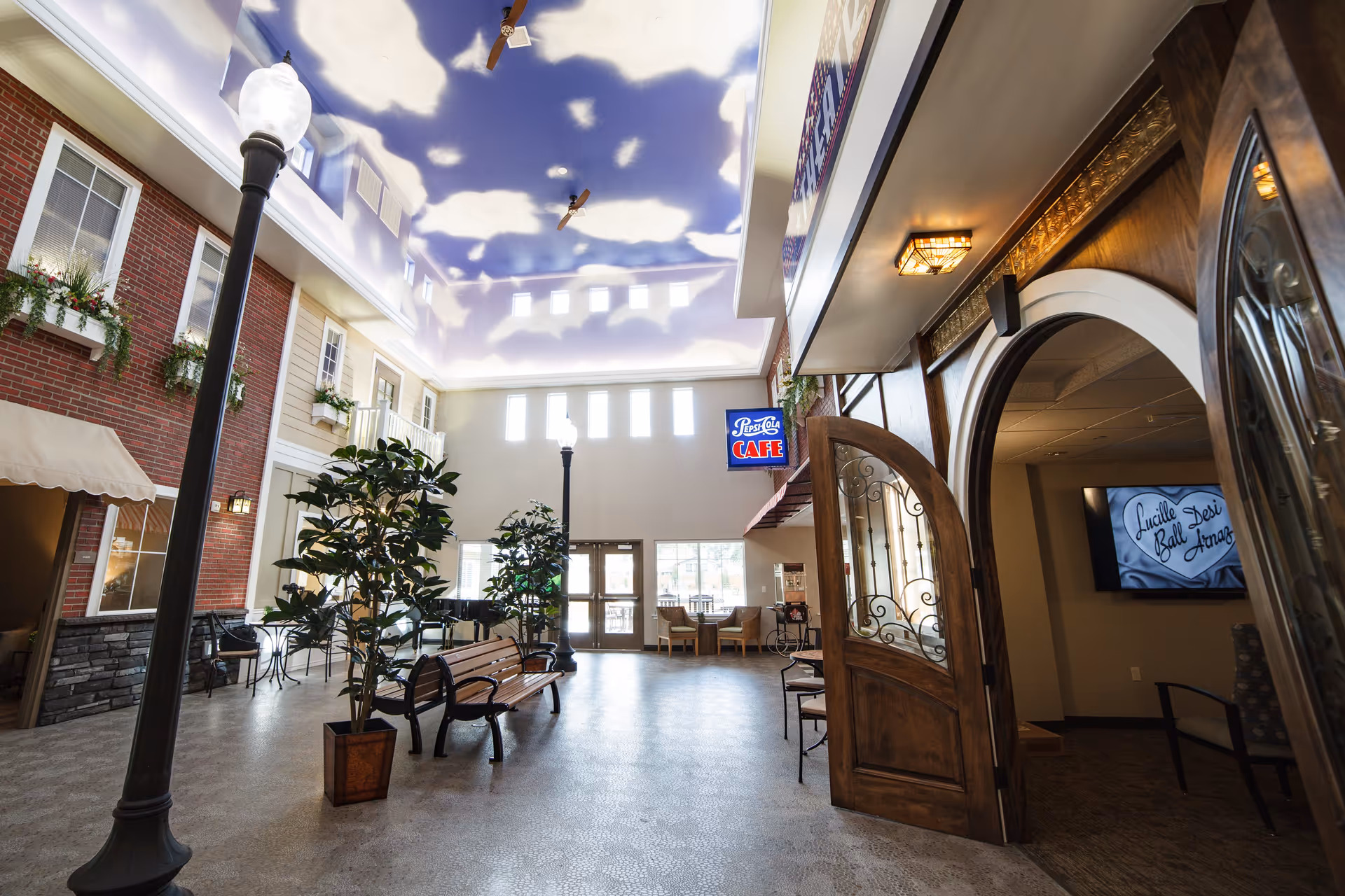 Indoor common area designed to resemble an outdoor street scene with a sky-painted ceiling featuring clouds and ceiling fans. The space includes street lamps, potted plants, benches, and a Pepsi-Cola Cafe sign. There are large windows and doors letting in natural light, and an open wooden door leading to another room with a TV screen displaying 'Lucille Desi Ball Arena'.