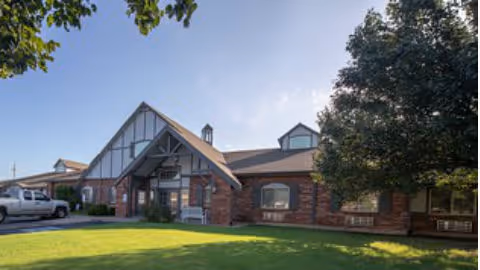 Exterior view of a single-story brick building with a gabled roof and large windows, surrounded by green grass and trees under a clear blue sky.