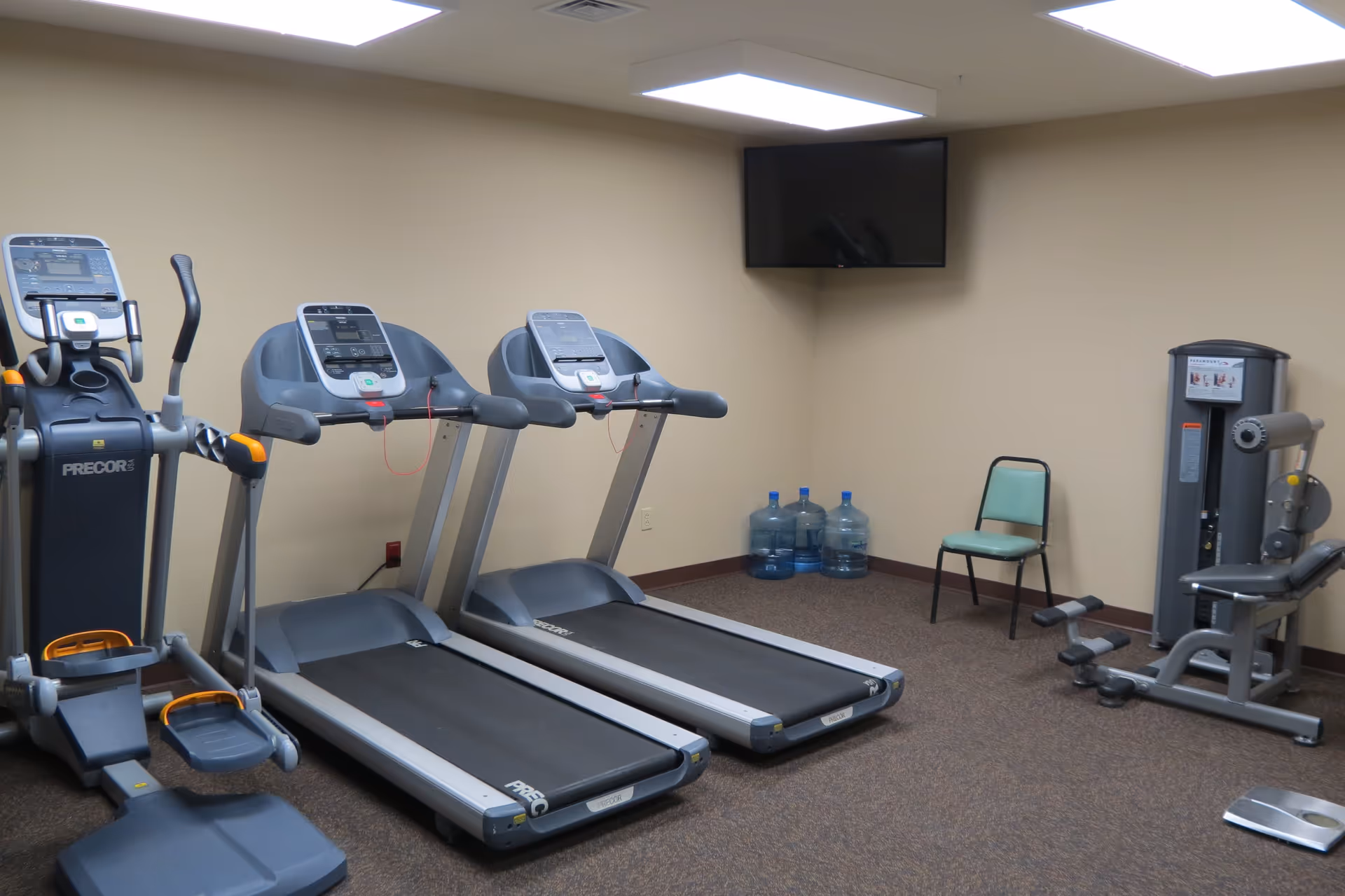 Indoor fitness room with two treadmills, an elliptical machine, a seated leg exercise machine, a green chair, three large water jugs in the corner, and a wall-mounted flat screen TV.
