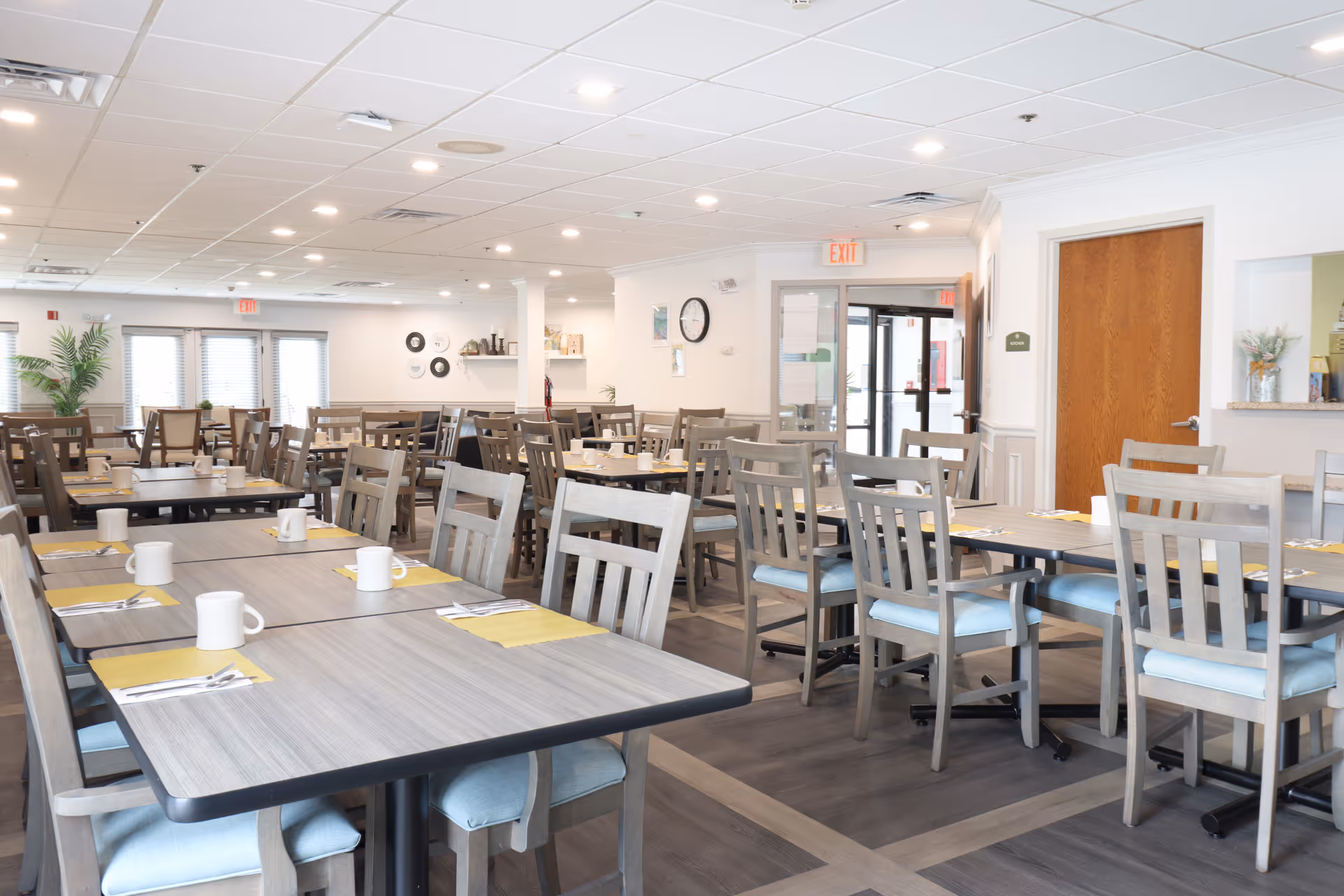 A bright and spacious dining room with multiple wooden tables and chairs arranged neatly. Each table is set with white mugs, yellow placemats, and silverware. The room has large windows with blinds, a clock on the wall, and light-colored walls and ceiling with recessed lighting.