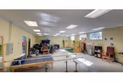 Bright interior therapy room with parallel bars, wheelchairs, exercise equipment and seating along the walls.