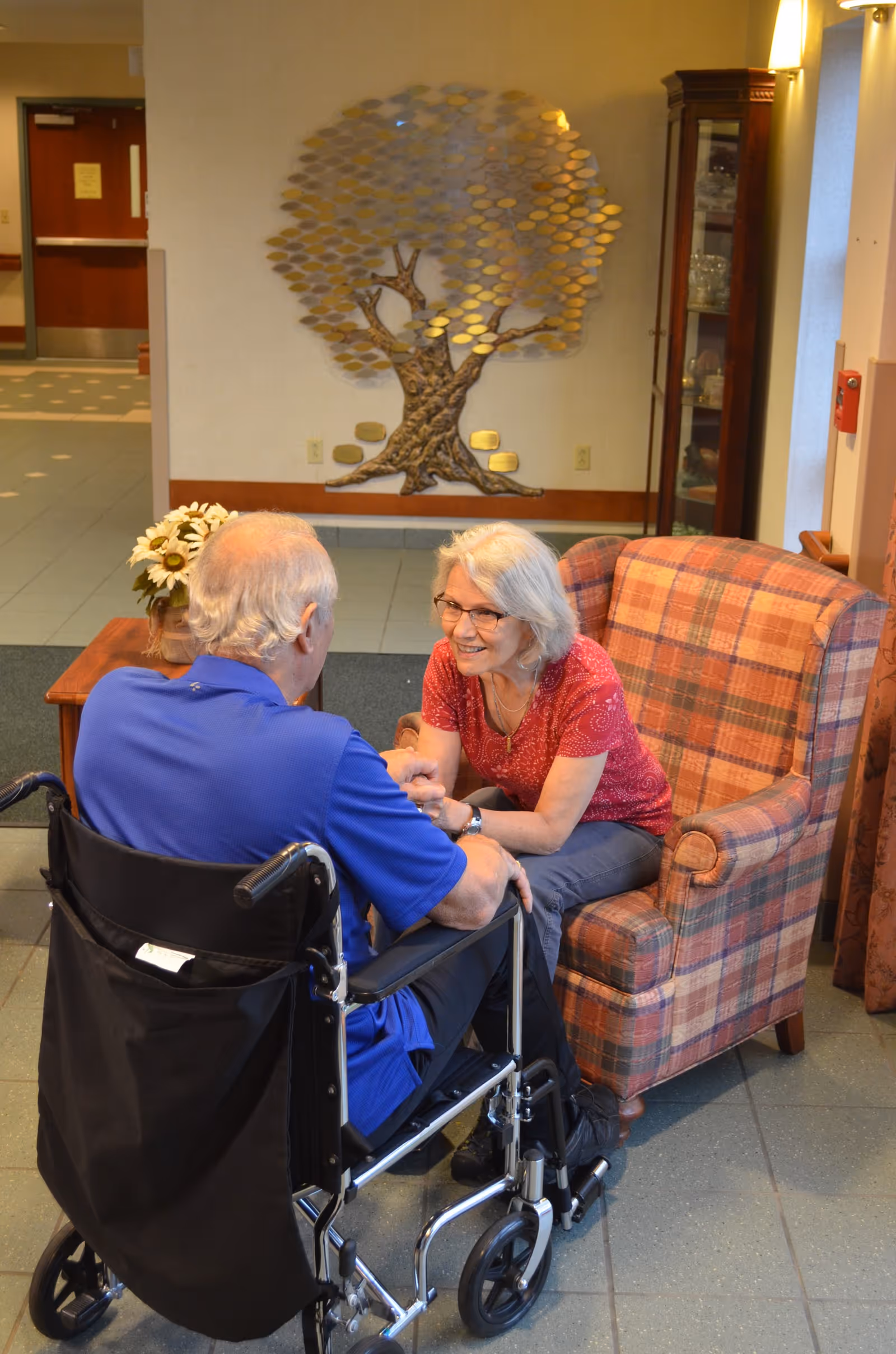 An elderly woman with gray hair and glasses sitting in a plaid armchair, holding hands and smiling at an elderly man in a wheelchair in a cozy indoor setting with a decorative tree wall art in the background.