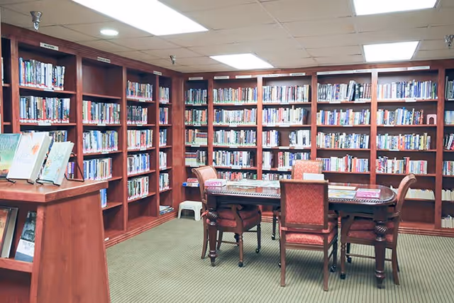 A cozy library room with wooden bookshelves filled with books lining the walls. In the center, there is a wooden table surrounded by four upholstered chairs. The room has a carpeted floor and a ceiling with recessed lighting.