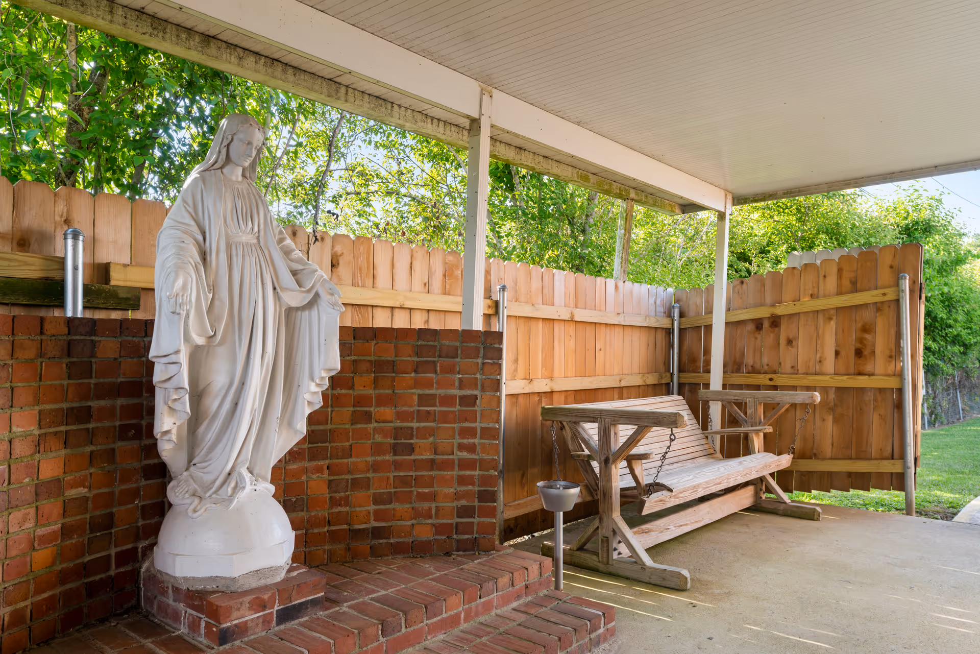 Covered outdoor patio showing a white religious statue on a brick base beside a wooden swing bench and wooden privacy fence.