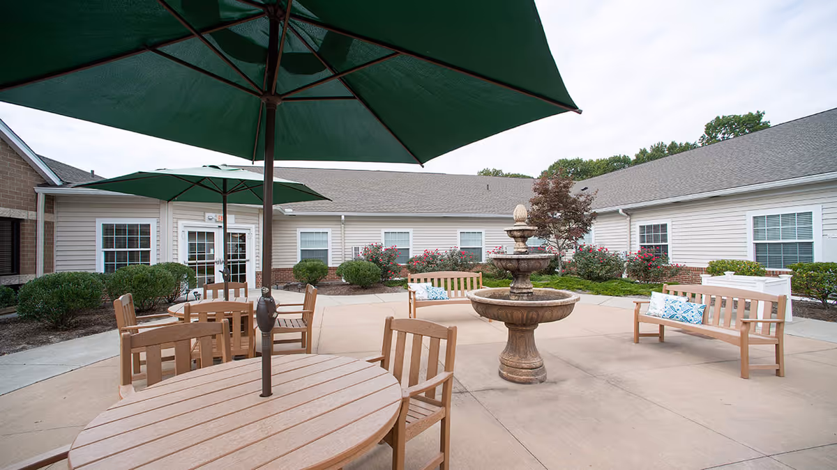 Outdoor courtyard area with wooden tables and chairs under large green umbrellas, wooden benches with cushions, a central stone fountain, and surrounding bushes and flowers in front of a single-story building with beige siding and white-framed windows.