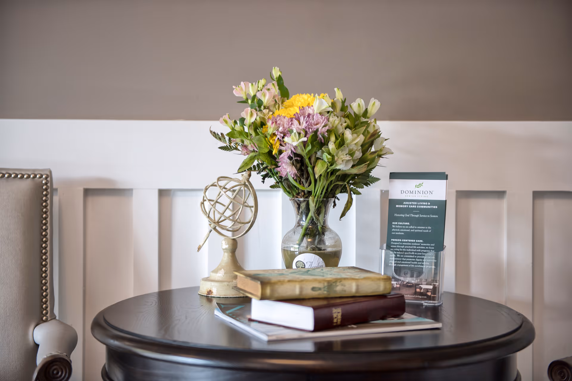 A round dark wooden table with a vase of colorful flowers, a decorative globe, two stacked books, and a brochure holder displaying information about Dominion Senior Living of Bristol. The background features a white paneled wall and part of a beige upholstered chair with nailhead trim.