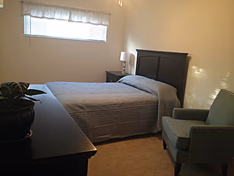 A simple bedroom with a bed covered in a gray quilt, a green upholstered armchair, a dark wooden dresser with a potted plant on top, a nightstand with a lamp, and a window with blinds and a white valance.