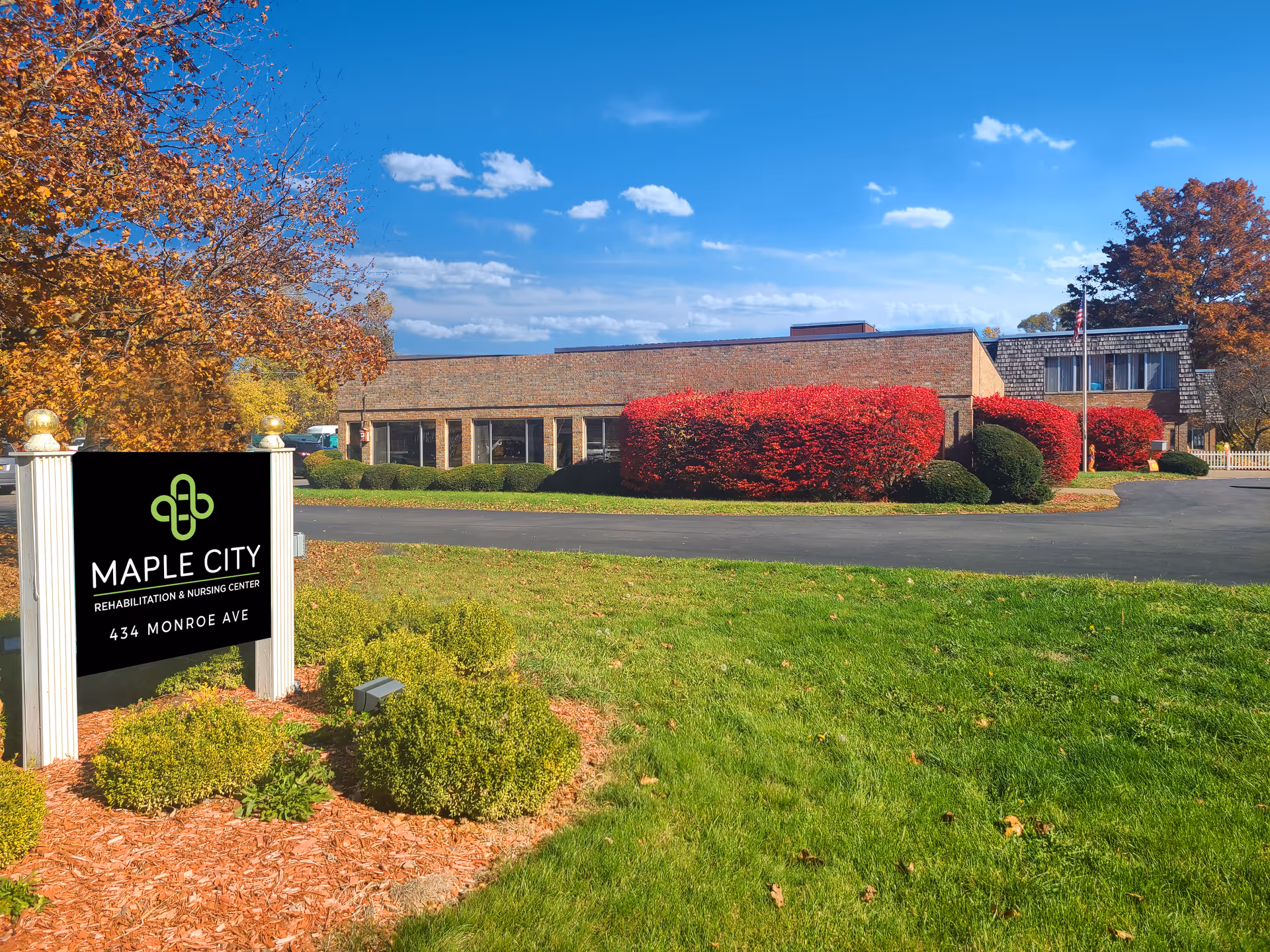 Exterior view of Maple City Rehabilitation and Nursing Center building with a clear blue sky, green lawn, and landscaped bushes including bright red shrubs. A black sign with white posts in the foreground displays the facility name and address: 434 Monroe Ave.