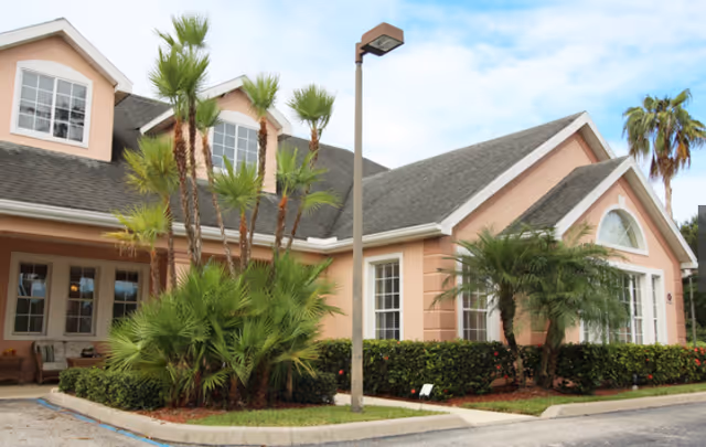 Front exterior of a peach-colored assisted living building with large windows, palm trees, a lamp post, and a small driveway.