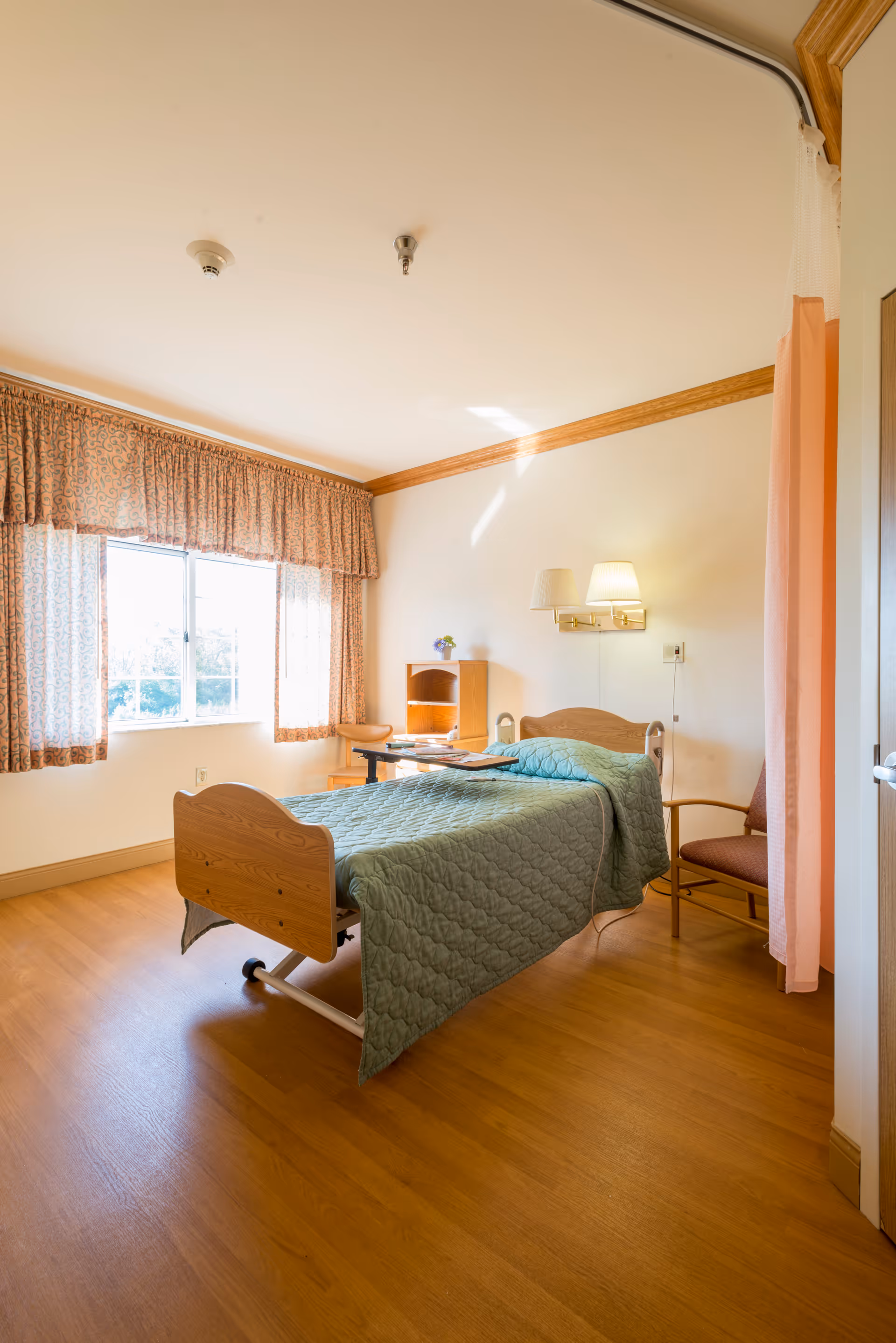 A bright and clean skilled nursing facility room with a single hospital bed covered in a green quilted blanket. The room has wooden flooring, a window with patterned curtains letting in natural light, a small wooden desk with a chair, a wall-mounted lamp, and a privacy curtain partially drawn to the side.