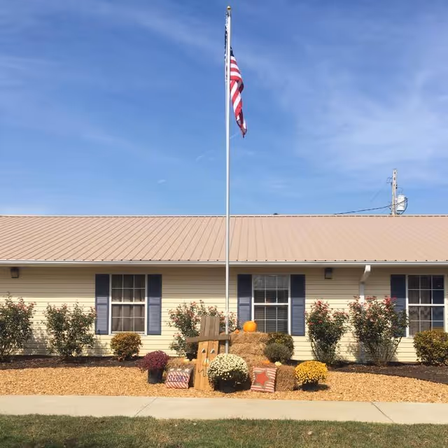 Single-story light-yellow building front with an American flag on a flagpole and fall decorations of hay bales, pumpkins, and flowers.
