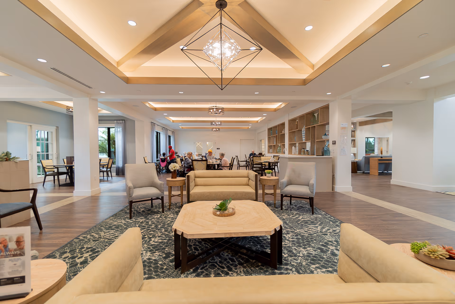 Open, well-lit senior living common area with sofas and armchairs around a central coffee table and dining tables with residents in the background.