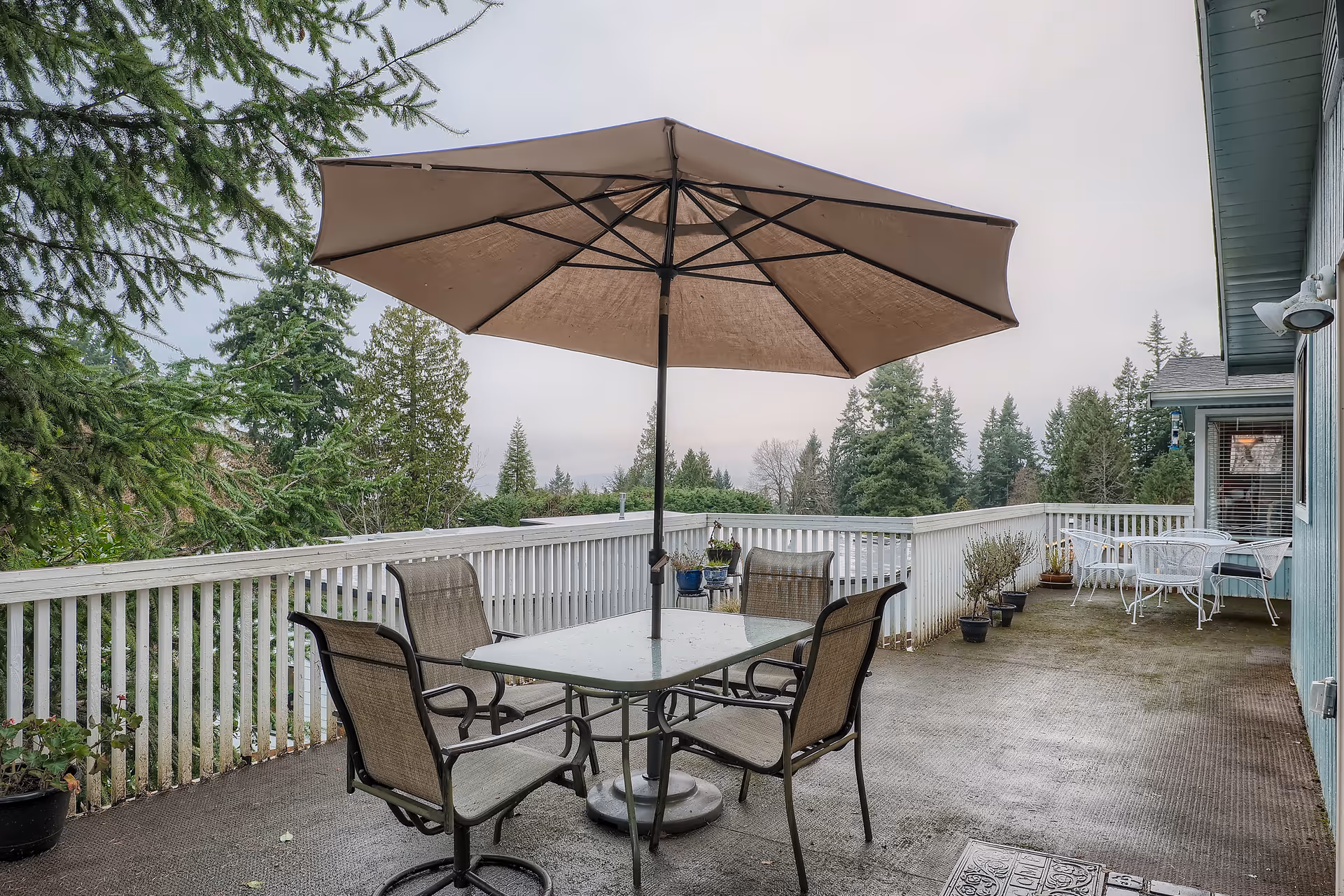 Outdoor patio area with a large beige umbrella over a glass table surrounded by four chairs. The patio has a white railing and several potted plants along the edge. In the background, there are tall evergreen trees and a cloudy sky. The side of a blue building with windows and outdoor lights is visible on the right.