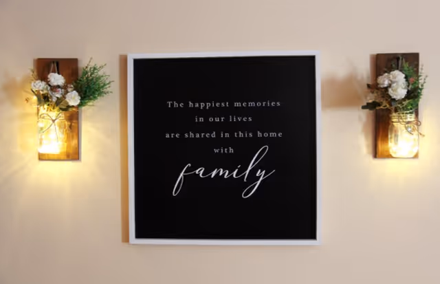 A wall decoration featuring a black framed sign with white text that reads 'The happiest memories in our lives are shared in this home with family', flanked by two wooden wall sconces with lit mason jar lights and white flowers with greenery.