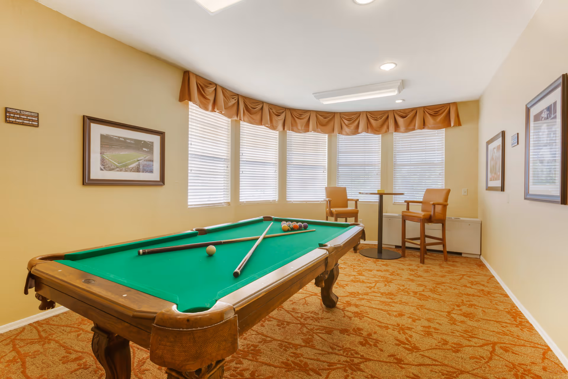 A room with a green felt pool table with two pool cues and billiard balls arranged on it. The room has beige walls, a patterned carpet, and a row of windows with blinds and a brown valance. There are two wooden chairs and a small round table near the windows, and framed pictures hang on the walls.