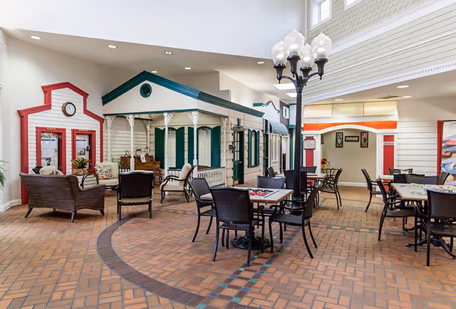 Indoor common area designed to resemble a small village street with house facades, featuring wicker chairs and tables with board games, a streetlamp-style light fixture, and a brick-patterned floor.