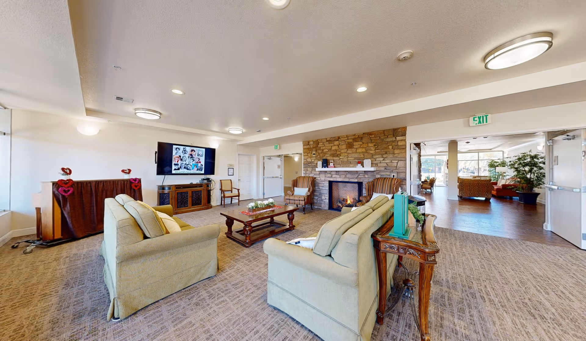A spacious living room area in a senior living facility with two beige sofas facing a wooden coffee table and a stone fireplace. There are two armchairs near the fireplace, a TV mounted on the wall, and a piano covered with a cloth decorated with red heart-shaped ornaments. The room has carpeted flooring and recessed lighting, with an open doorway leading to another seating area with large windows and wooden flooring.