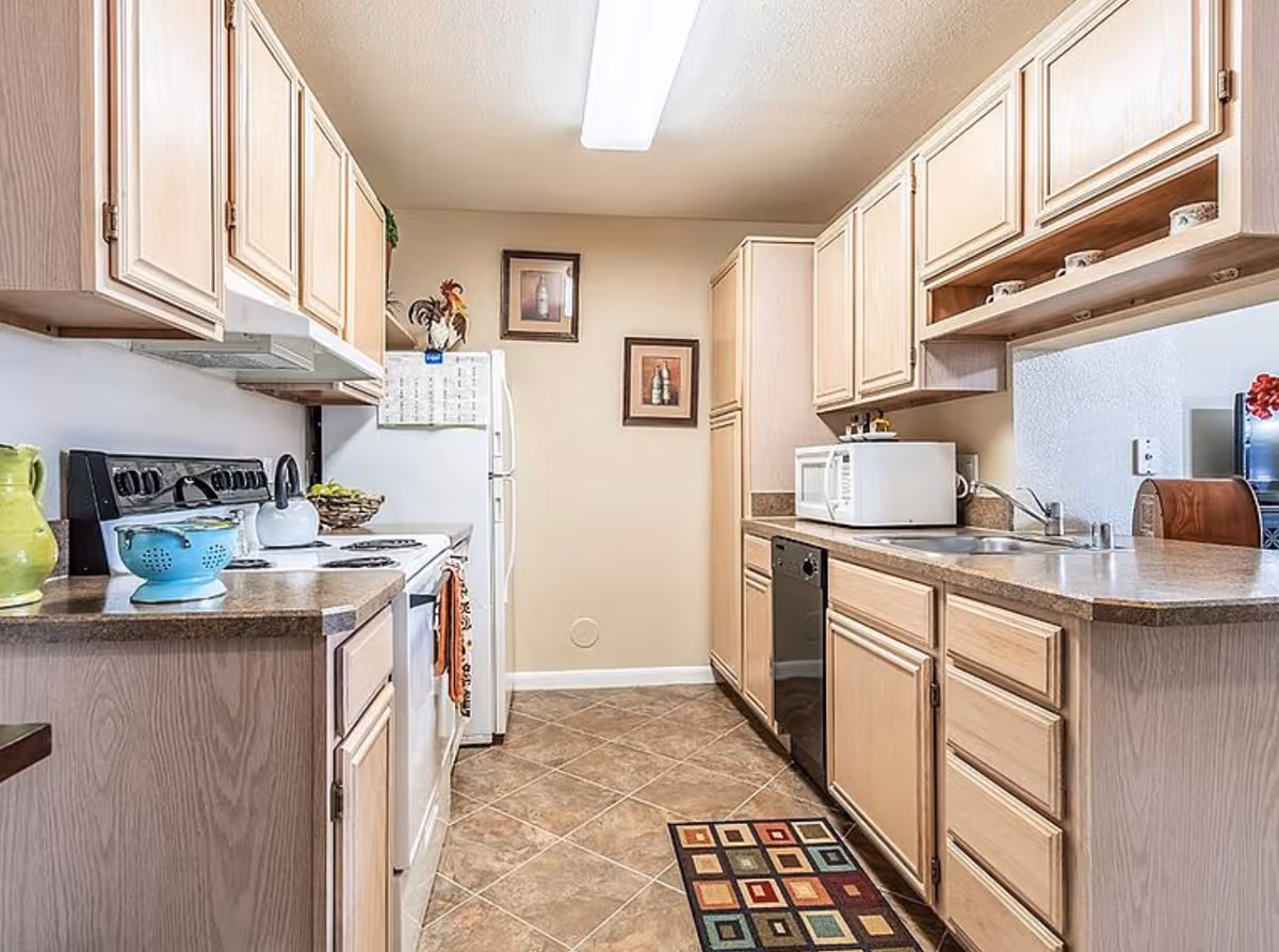 A bright kitchen with light wood cabinets, a white refrigerator, stove, microwave, dishwasher, and a double sink. The floor is tiled with a small colorful rug in front of the sink. There are two framed pictures on the far wall and various kitchen items on the countertops.