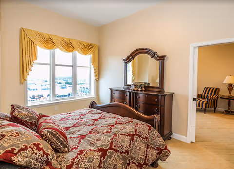 A bedroom with a large bed covered in a red and beige patterned comforter and matching pillows. There is a wooden dresser with a mirror against the wall opposite the bed. A window with yellow drapes lets in natural light. An open doorway leads to another room with a striped armchair and a side table with a lamp.