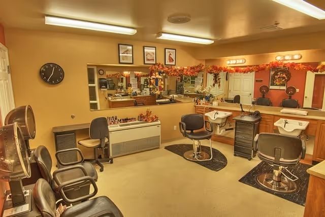 Interior view of a salon area in a senior living facility with multiple salon chairs, hair dryers, mirrors, and wash basins. The room is decorated with autumn-themed garlands above the mirrors and has framed pictures on the wall. There is a clock on the wall and various salon equipment and supplies are visible.
