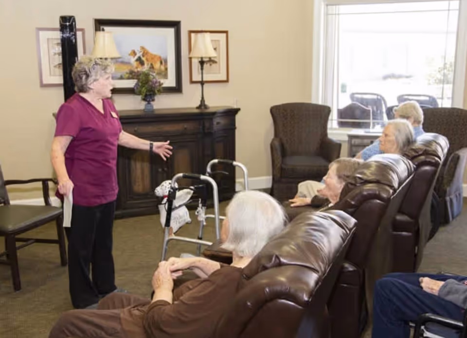 A woman in a maroon uniform stands and gestures while speaking to a group of elderly people seated in leather recliners in a cozy living room setting with armchairs, a window, and framed artwork on the wall.
