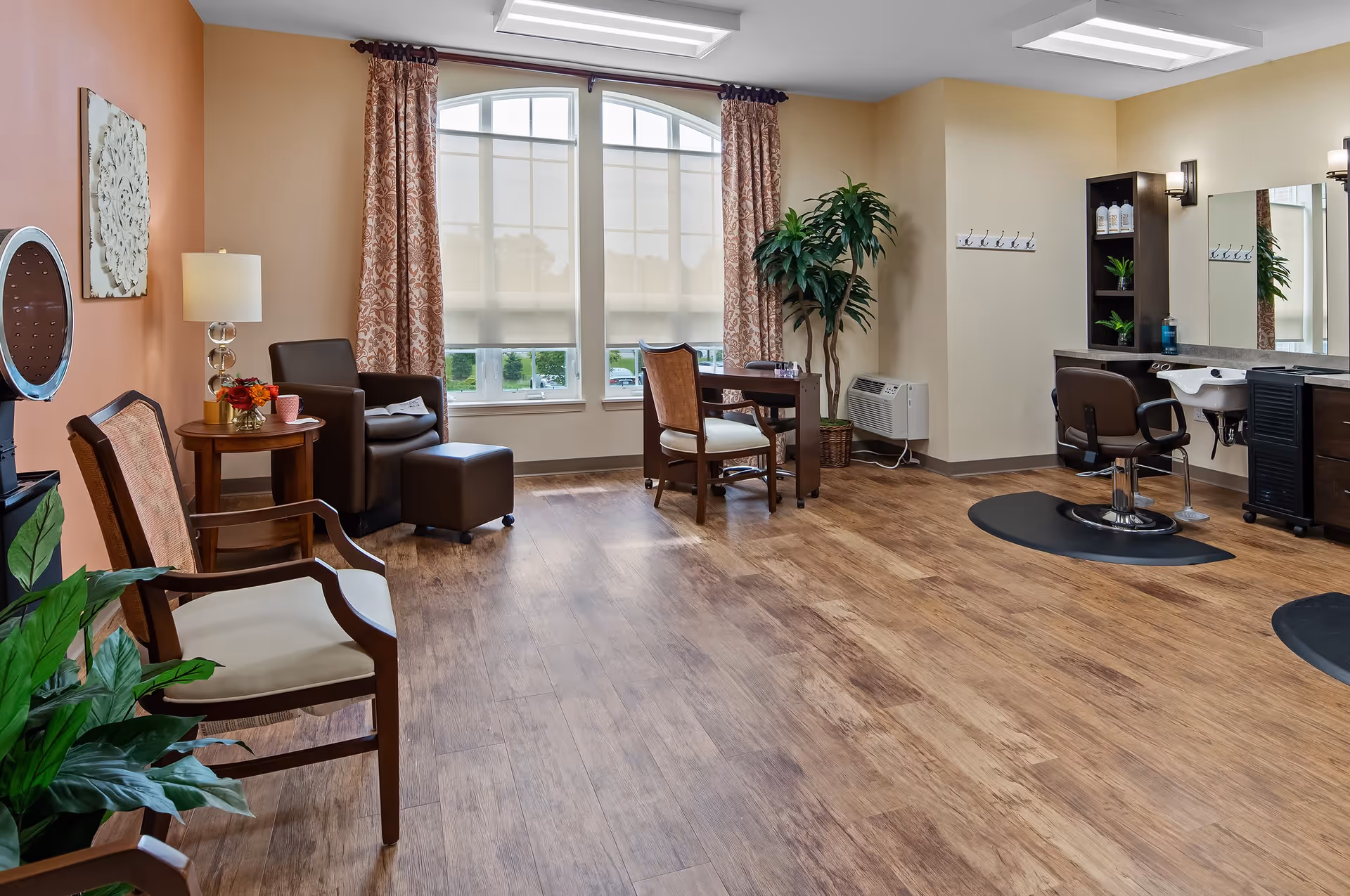 A bright and spacious salon area with wooden flooring, featuring a styling chair and sink station on the right, a small table with a chair in front of large windows with patterned curtains, a comfortable armchair with an ottoman, a side table with a lamp and flowers, and decorative plants.