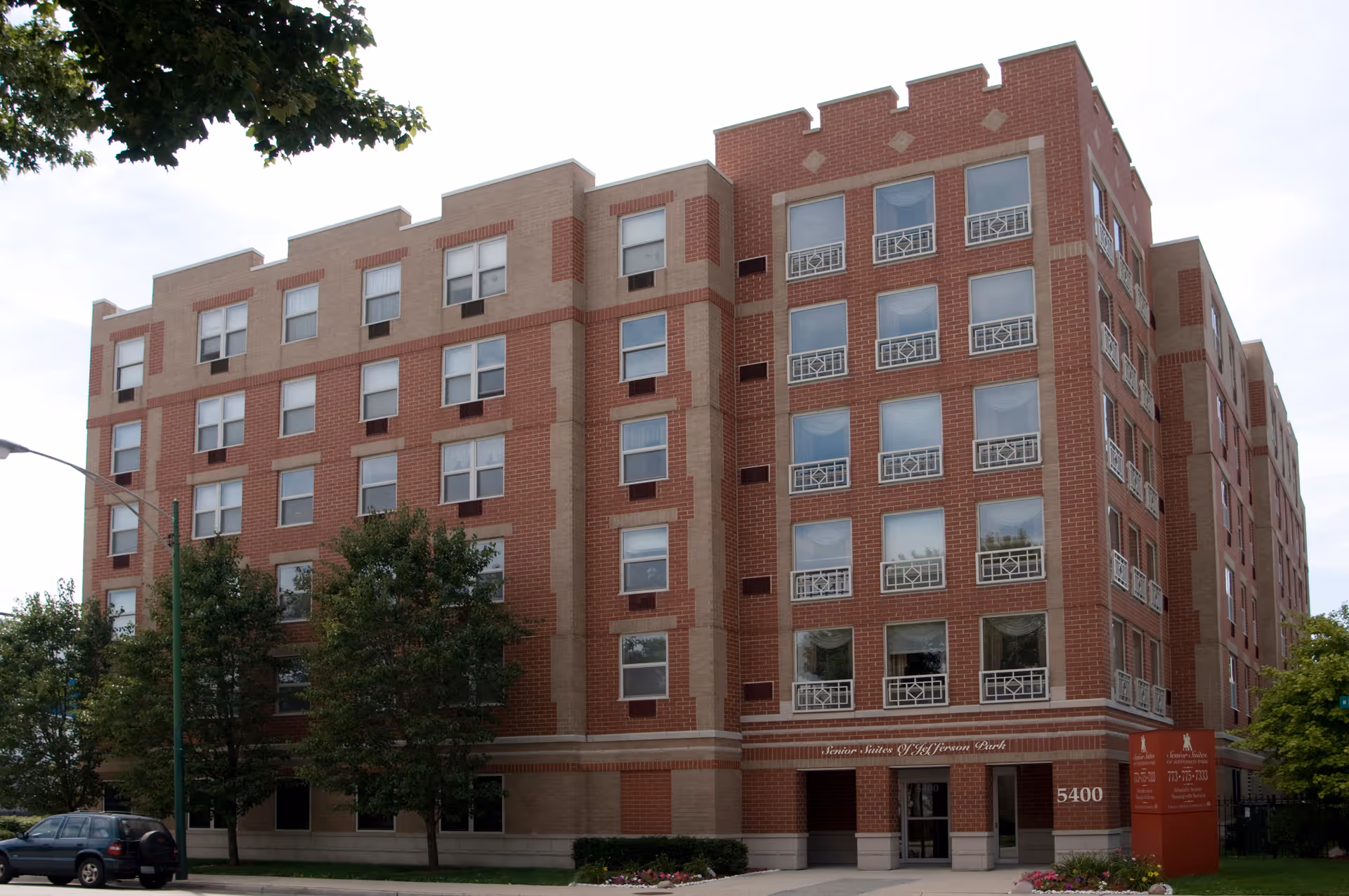 Exterior view of a multi-story brick building with numerous windows, trees in front, and a sidewalk leading to the entrance. The building has a sign near the entrance that reads 'Senior Suites of Jefferson Park'.