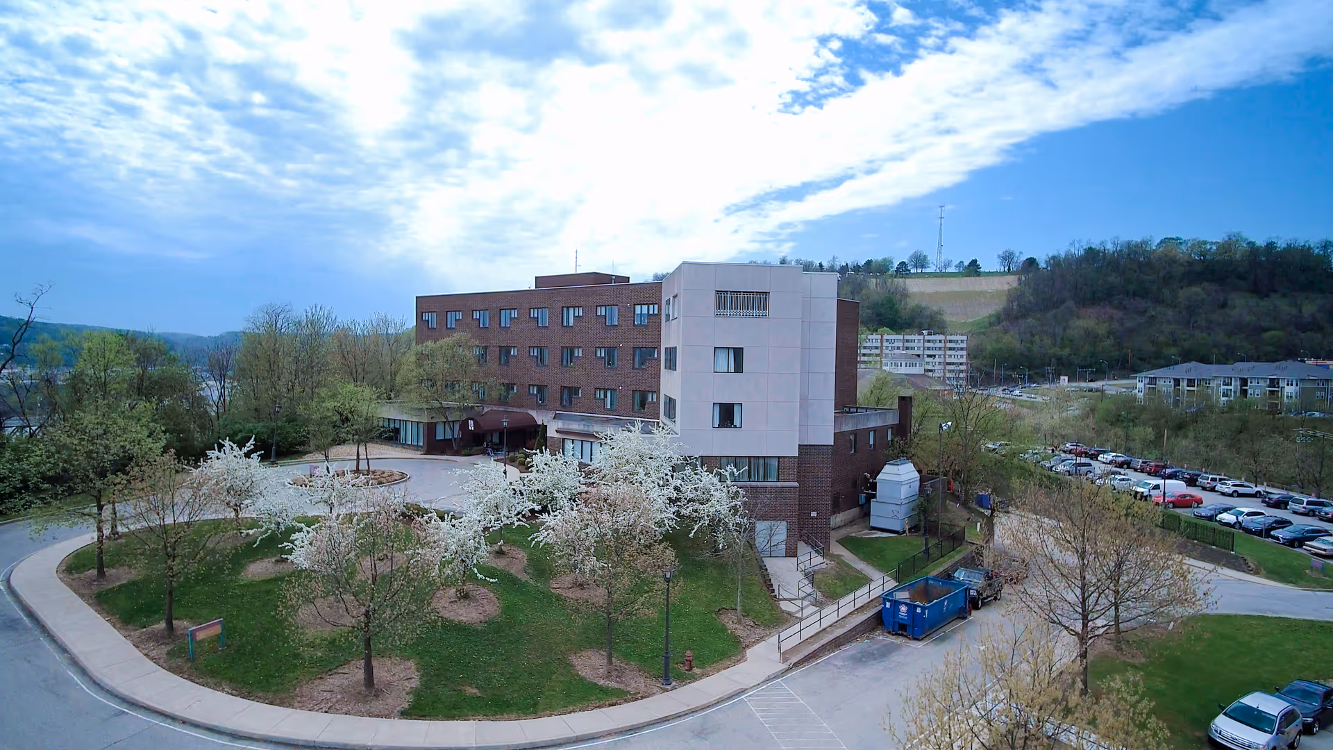 Exterior view of a multi-story senior living facility building surrounded by trees and greenery with a curved driveway and parking area. The sky is partly cloudy with blue patches visible.
