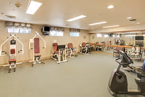 A spacious fitness room with various exercise machines including weight machines, stationary bikes, and other gym equipment. The room has beige walls, a green carpeted floor, small windows with valances, and ceiling lights.