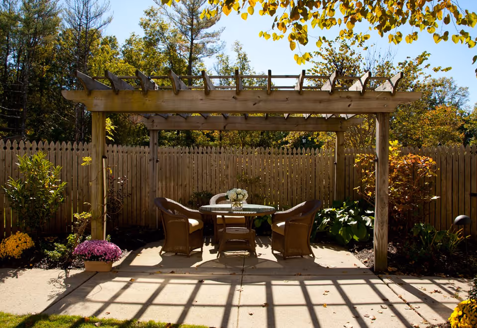 Outdoor seating area with a wooden pergola casting shadows on a concrete patio. There is a round glass table with a flower arrangement in the center and four wicker chairs around it. The area is enclosed by a wooden picket fence and surrounded by various plants and trees with autumn foliage.