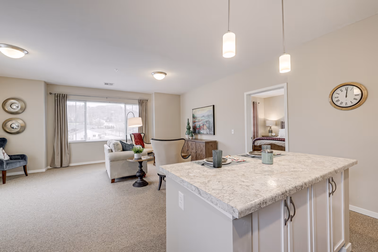 Open-plan living area with a kitchen island in the foreground, a seating area by a large window, and a bedroom visible through a doorway.