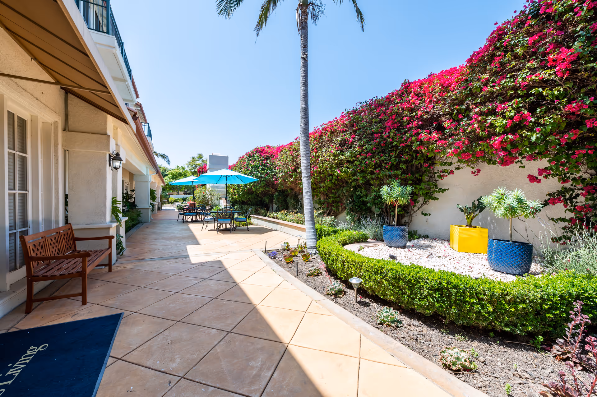 Outdoor patio area at Aegis Living Granada Hills featuring a tiled walkway with a wooden bench on the left, a garden bed with green shrubs and colorful potted plants on the right, bright pink bougainvillea climbing a wall, and several tables with blue umbrellas in the background under a clear blue sky.