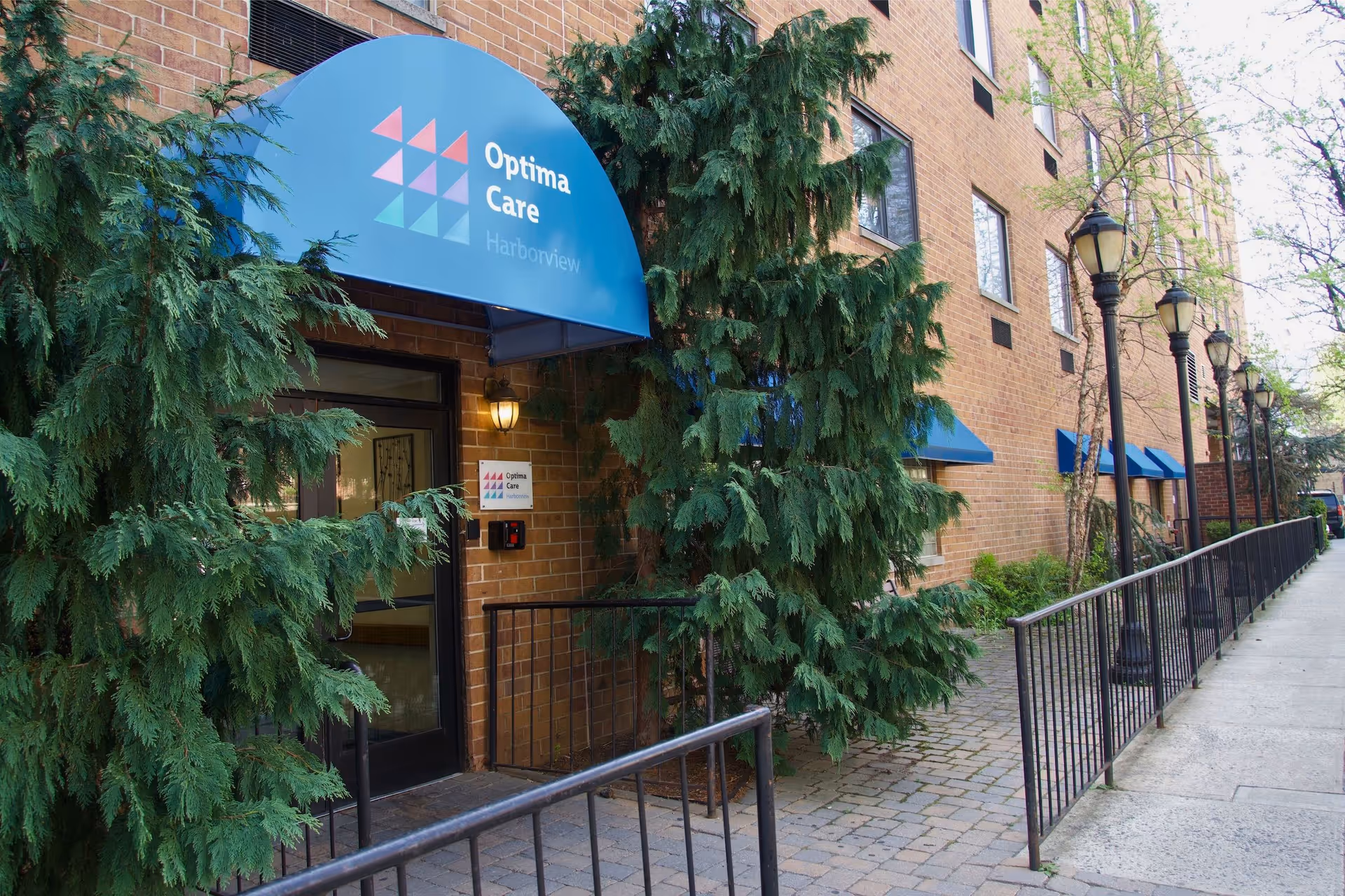 Entrance of Optima Care Harborview showing a brick building with a blue awning, evergreen trees, a railing and a sidewalk lined with lampposts.