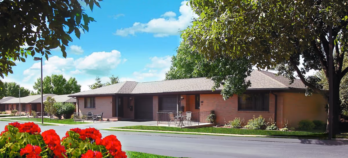 Single-story brick residential buildings with attached garages, small patios with chairs, surrounded by green lawns, trees, and bright red flowers in the foreground under a blue sky with scattered clouds.