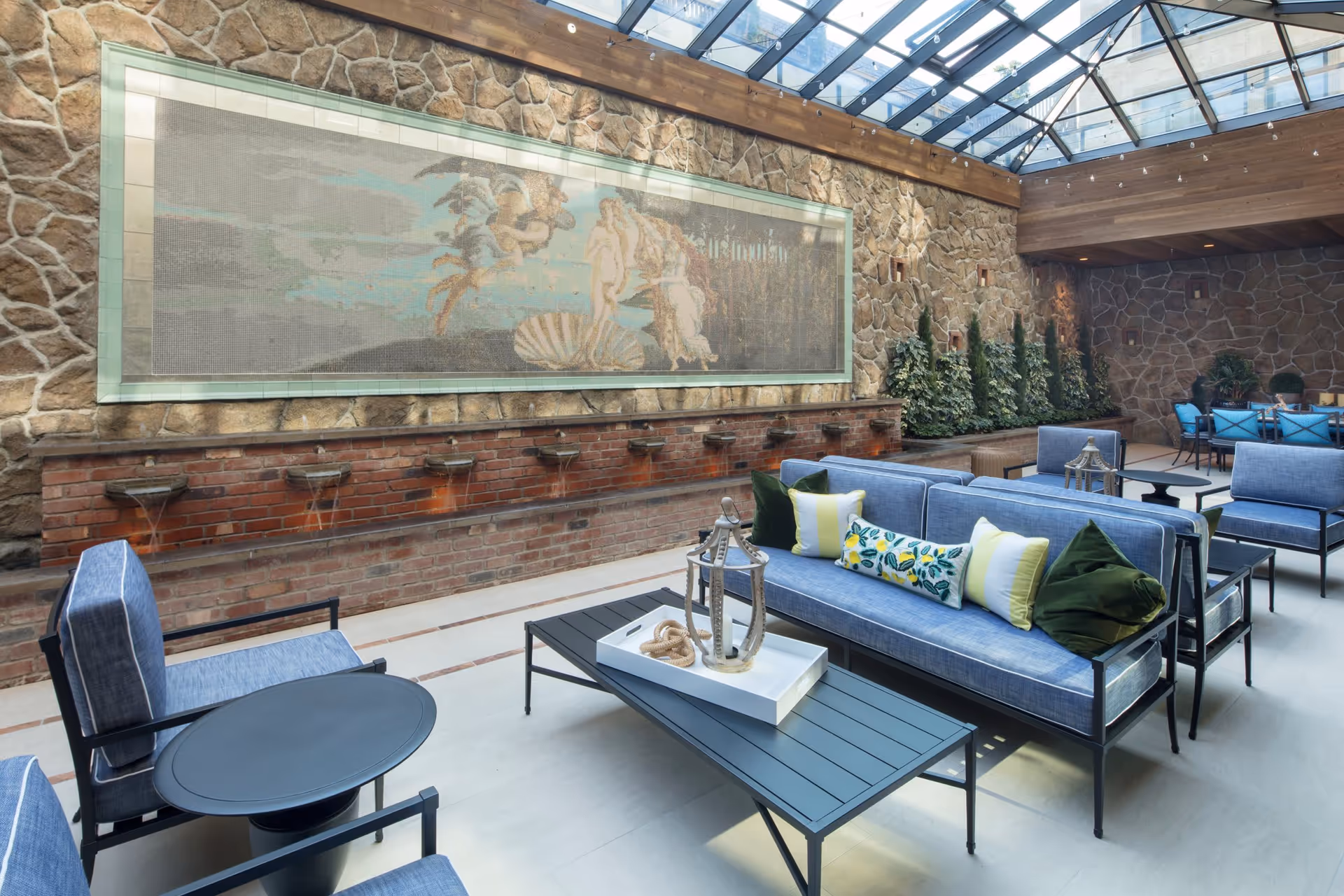 Indoor lounge area with blue cushioned seating, a black coffee table with decorative items, stone walls, a large mosaic artwork on the wall, and a glass ceiling allowing natural light to fill the space.
