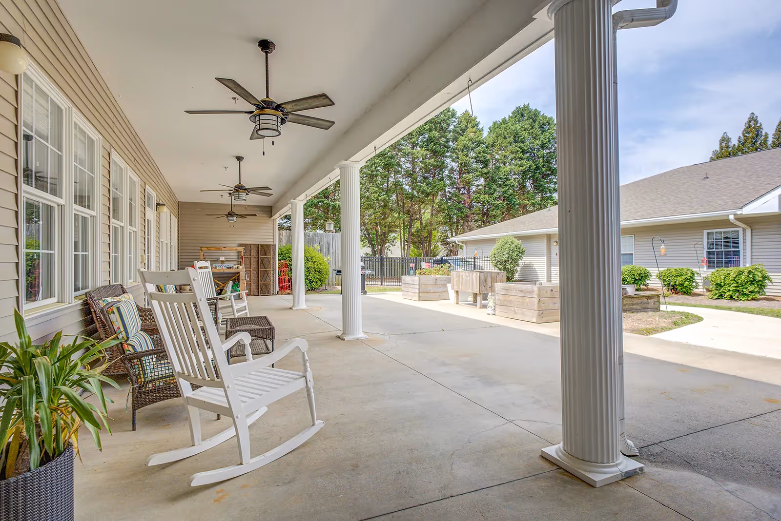 Covered outdoor patio area with white rocking chairs, wicker chairs with cushions, ceiling fans, and potted plants. The patio overlooks a garden area with raised wooden planters and trees in the background.